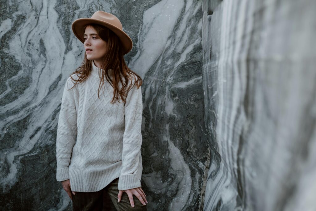 Woman in White Knitted Sweater and Brown Fedora Hat Standing Beside Gray Marble Wall