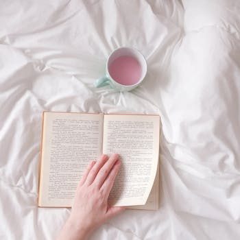 Relaxing scene of a hand turning book pages with a cup on white bedding, perfect for cozy reading vibes.