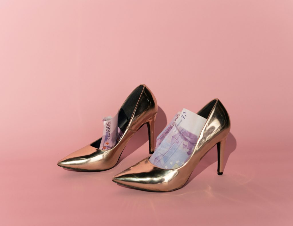 Stylish golden high heels stuffed with euro banknotes on a pink background, symbolizing luxury and wealth.